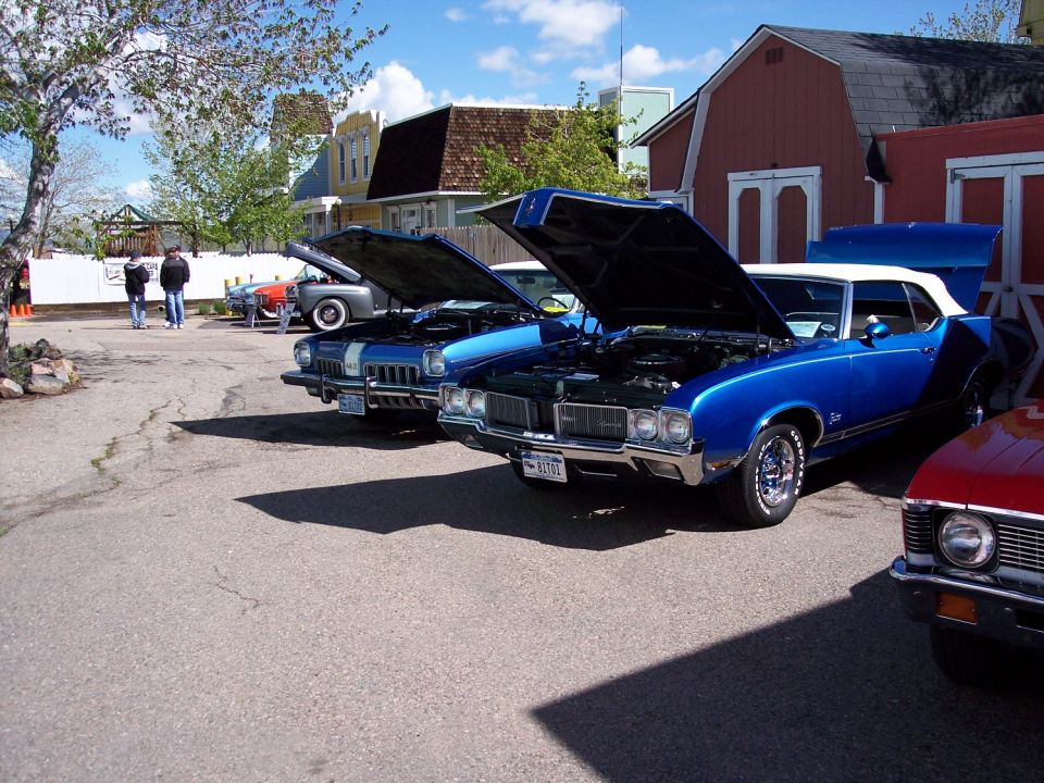 Three classic cars parked outside with hoods up. The closest car is blue with a white top