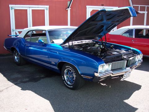 1972 Ford Ranchero - A blue convertible with its hood open is parked in front of a red building, with another car visible behind it.
