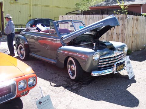 1972 Ford Ranchero - A gray convertible with its hood open sits parked next to an orange car. The vehicle has whitewall tires and a prominent chrome grille.