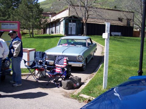 1972 Ford Ranchero - A light blue coupe is parked in front of a building with a grassy area and trees. Two chairs and bags are set up nearby.