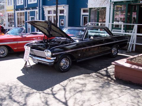1972 Ford Ranchero - A black 1963 Chevrolet Nova coupe is displayed with its hood open, parked alongside a red convertible in front of buildings.