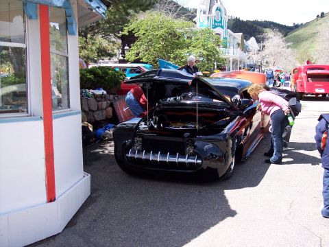 1972 Ford Ranchero - A crowd gathers around a custom black hot rod with its hood raised, revealing the engine compartment.