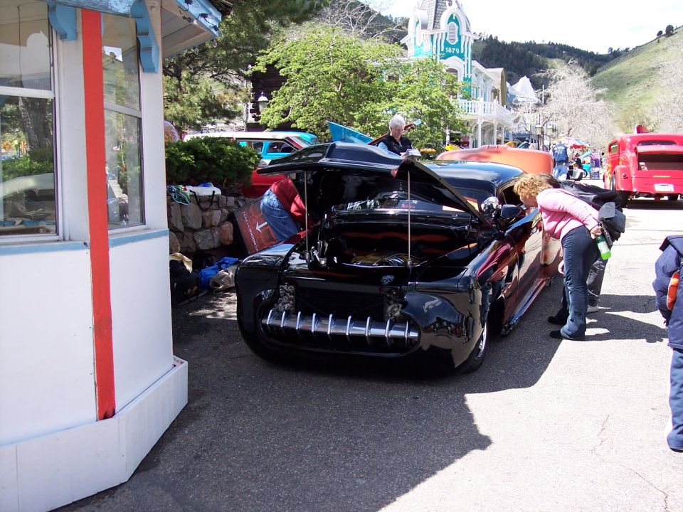 Black custom car with hood open, people looking at engine. Other vehicles and buildings in background
