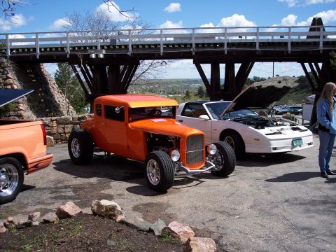 1972 Ford Ranchero - An orange hot rod coupe is parked next to a white car with its hood open under a bridge.