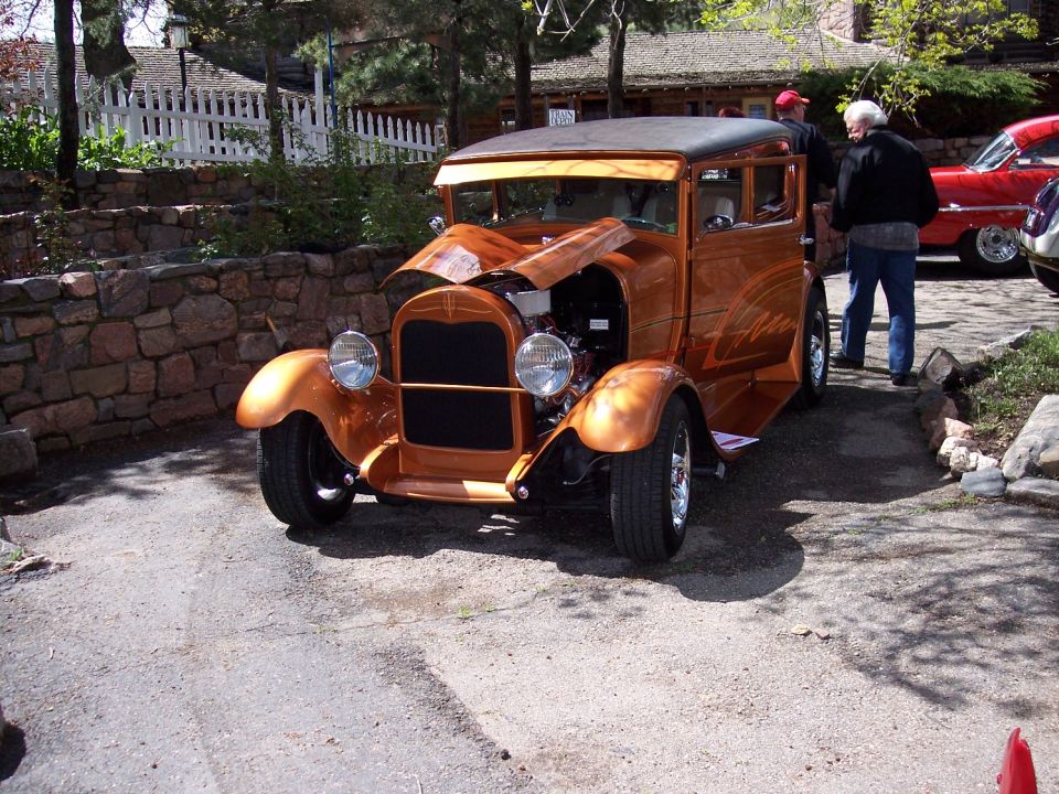 Copper-colored hot rod sedan with hood open, parked outside. Another red car is visible to the right