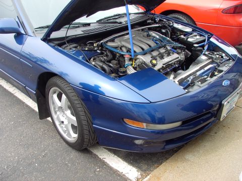 1989 Ford Probe - The engine compartment of a blue car is displayed with the hood raised, while a red vehicle is partially visible in the background.