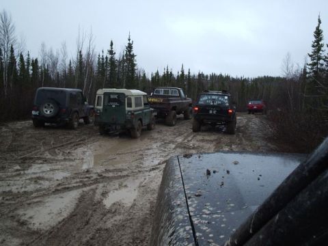 1986 Ford F-150 - Multiple off-road vehicles navigate a challenging muddy path in a wooded area.