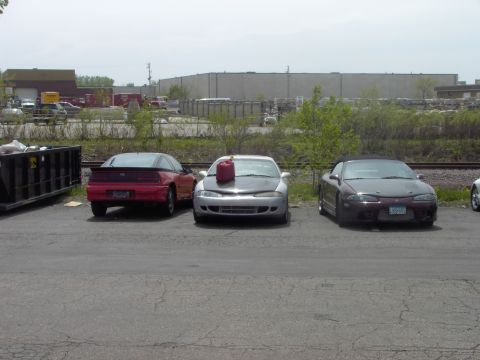 1989 Ford Probe - Three coupes are parked in an industrial lot with a dumpster and buildings in the background.