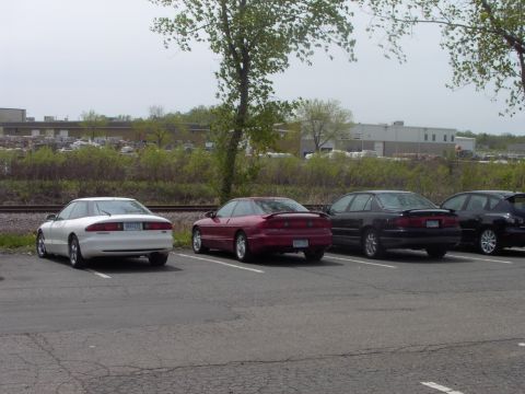 1989 Ford Probe - Multiple Ford Probes and a sedan parked in a lot with industrial buildings in the background.