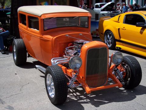 1972 Ford Ranchero - A bright orange hot rod coupe with a tan roof and exposed chrome engine sits parked next to a yellow modern sports car.