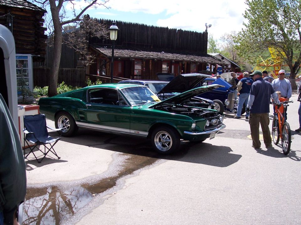 Green 1967 Ford Mustang Fastback with hood open, parked outside wooden buildings