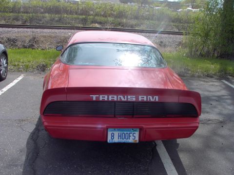 1989 Ford Probe - The rear of a red Pontiac Trans Am is visible, parked in a lot with railroad tracks in the background.
