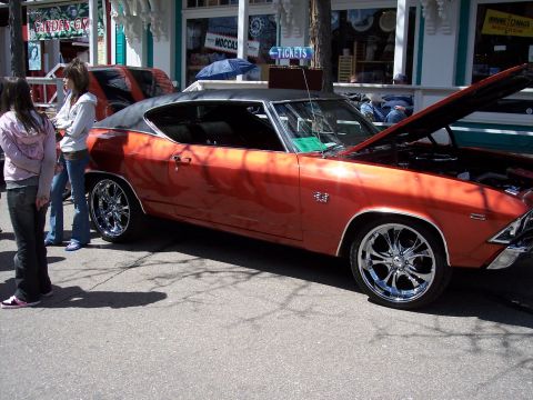 1972 Ford Ranchero - A classic orange muscle car with a black vinyl top and custom chrome wheels is parked on a street with people nearby.