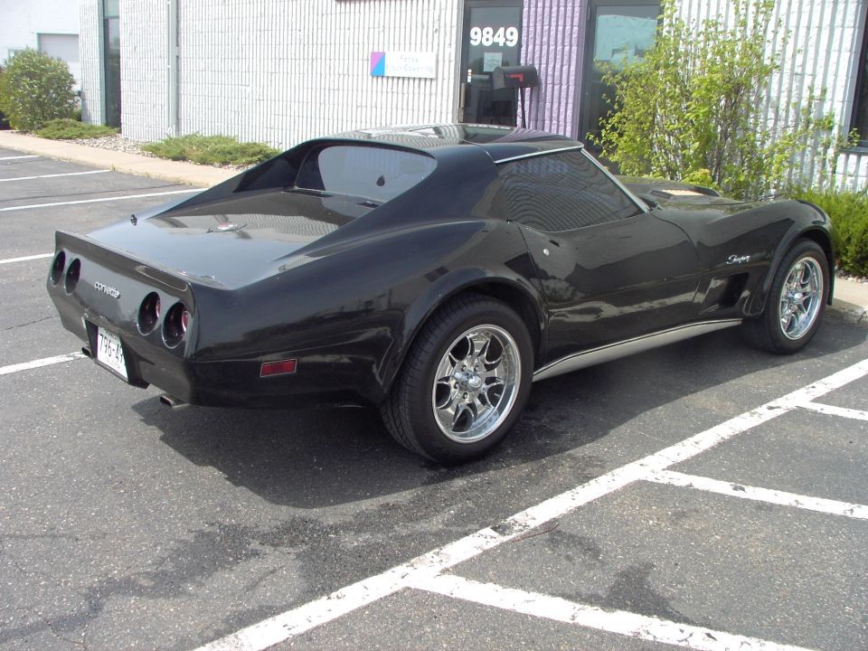 Black Corvette parked in a lot, rear view. Has chrome wheels and a spoiler