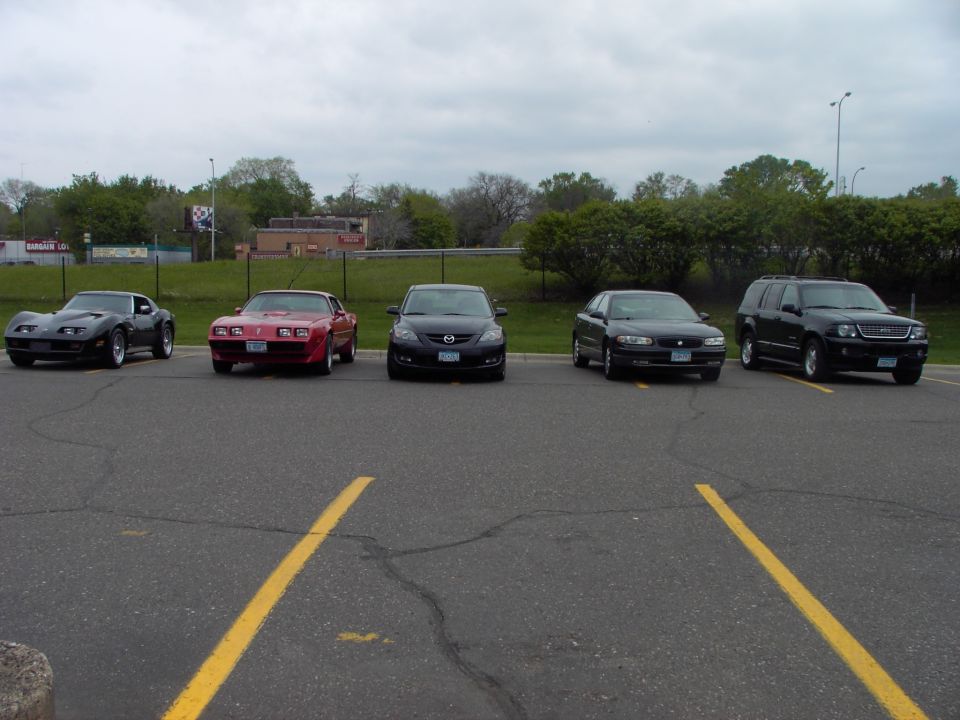 Five cars parked in a lot: black Corvette, red Pontiac Firebird, black Mazda, black sedan, black SUV