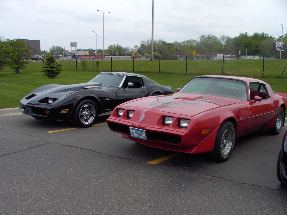 Red 1980s Pontiac Firebird with hood decal and black Chevrolet Corvette parked side-by-side