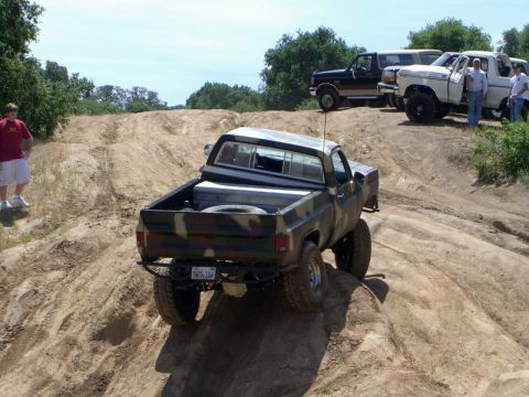 1980 Ford F-250 - A camouflage pickup truck navigates a challenging off-road course, with other vehicles parked on a ridge above.