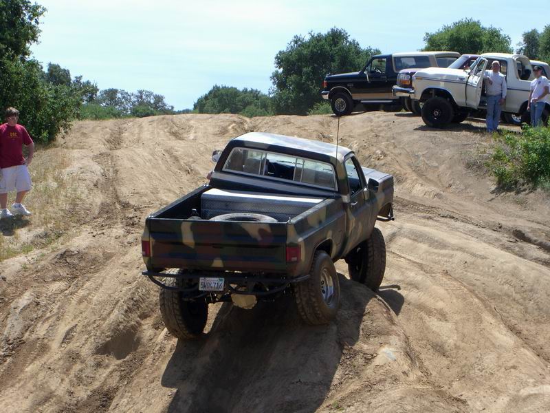 Rear view of camouflage pickup truck with lifted suspension on dirt trail. Other trucks visible in background
