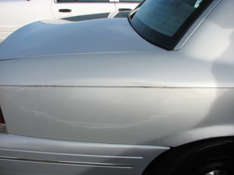 2003 Ford Crown Victoria - A silver 2003 Ford Crown Victoria is seen from the rear quarter panel, with another white vehicle partially visible in the background.