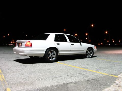 2003 Ford Crown Victoria - White Ford Crown Victoria sedan photographed at night from the passenger-side rear three-quarter in an empty parking lot, with the tailligh…