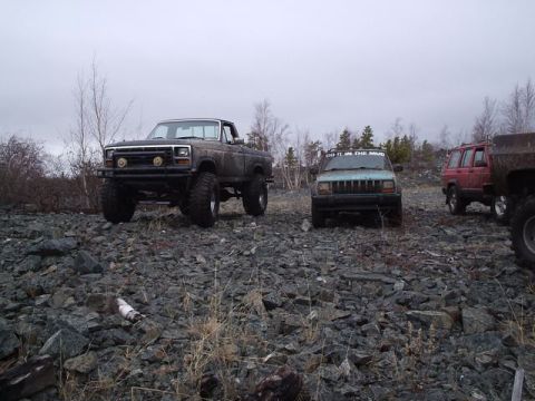 1986 Ford F-150 - A lifted brown pickup truck is parked on a rocky terrain, with other off-road vehicles visible in the background.