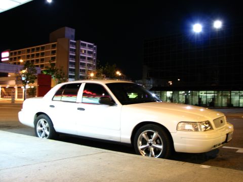 2003 Ford Crown Victoria - Night shot of a white Ford Crown Victoria sedan parked at a downtown curb, seen from a low three-quarter front angle with polished five-spo…