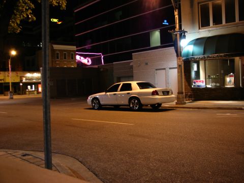 2003 Ford Crown Victoria - A white 2003 Ford Crown Victoria is parked on a city street at night, with buildings in the background.