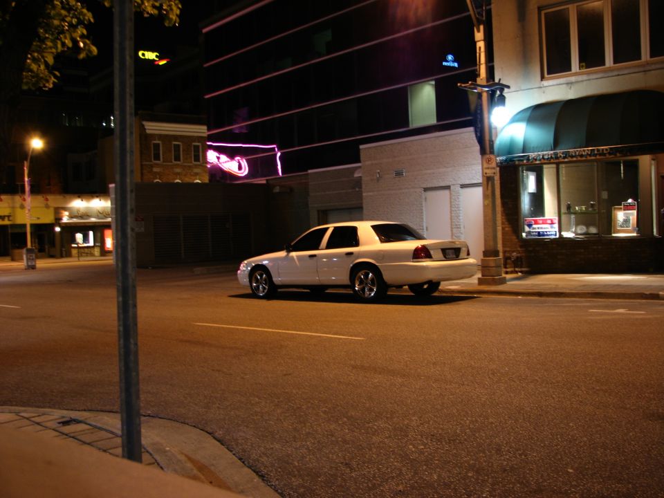White 2003 Ford Crown Victoria parked on a city street at night. The car is seen from the rear