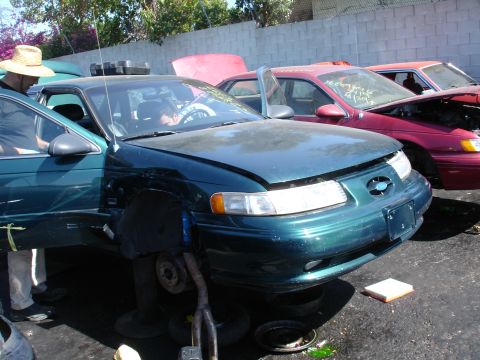1993 Ford Taurus - Junkyard scene with a green Ford Taurus and other vehicles, one with its hood open.