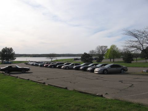 1988 Mercury Grand Marquis - Wide shot of a car-club meet in a lakeside park parking lot, with a long row of cars angle-parked facing the water, a black Firebird at lef…