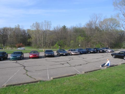 1988 Mercury Grand Marquis - A group of cars are parked in a lot with trees in the background and a person sitting on the curb.