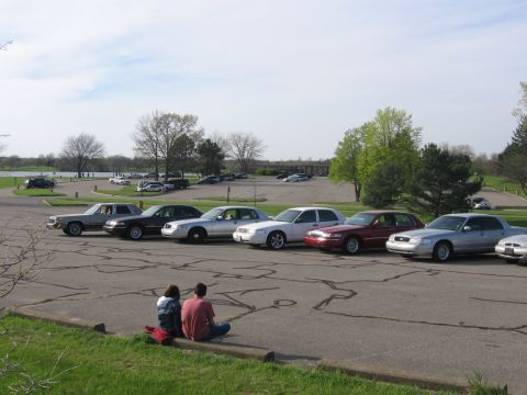 1988 Mercury Grand Marquis - A group of sedans are parked in a lot, with two people sitting in the foreground.