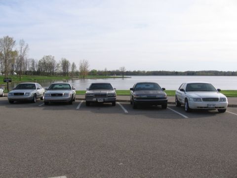 1988 Mercury Grand Marquis - Several sedans are parked in a lot next to a body of water on a cloudy day.