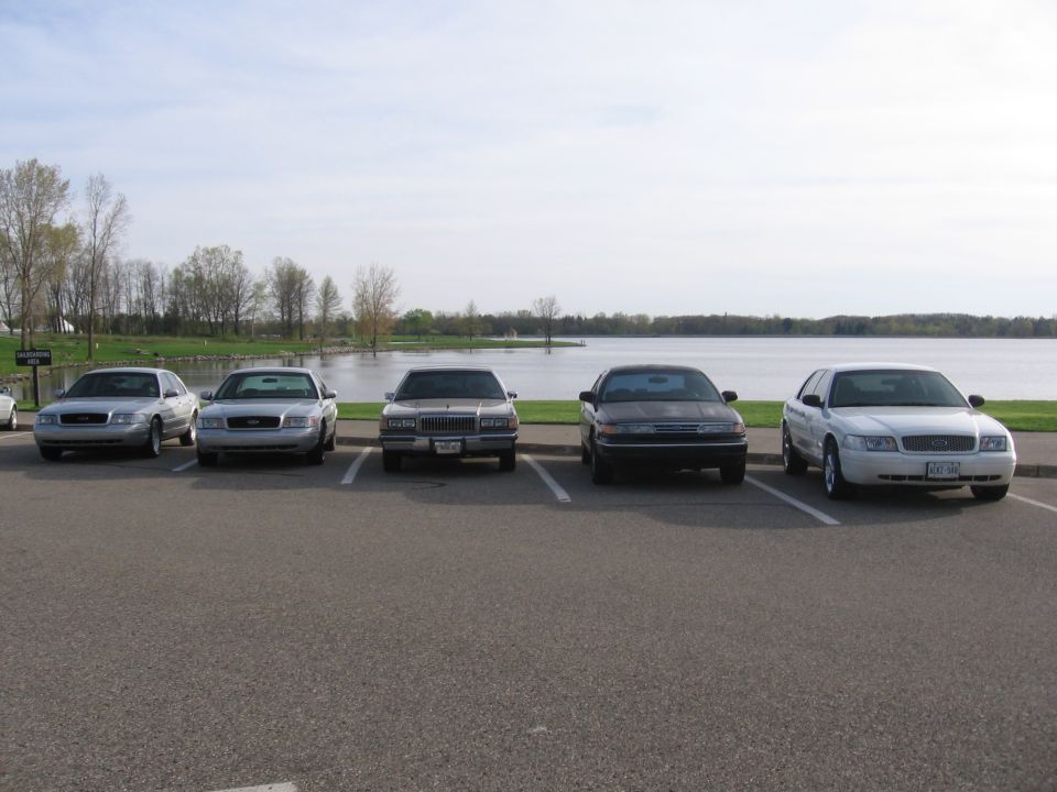 Five sedans parked in a row facing a lake; the middle car appears to be a 1998 Mercury Grand Marquis
