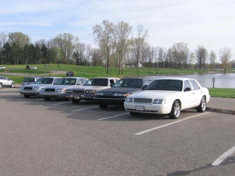 1988 Mercury Grand Marquis - Several sedans are parked in a lot near a body of water, with the white Ford Crown Victoria in the foreground.