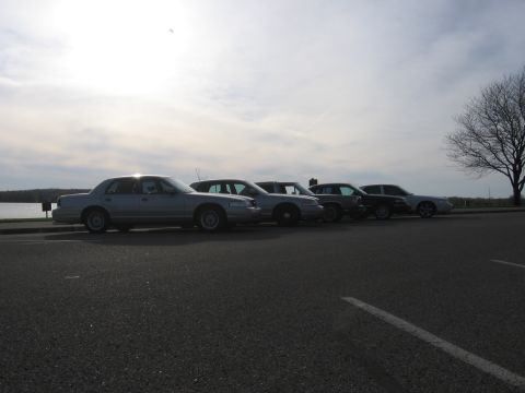 1988 Mercury Grand Marquis - A line of four sedans, including a silver 1988 Mercury Grand Marquis, are parked near a body of water under a cloudy sky.