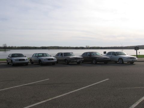 1988 Mercury Grand Marquis - Several sedans are parked in a lot with a lake and trees visible behind them under a cloudy sky.