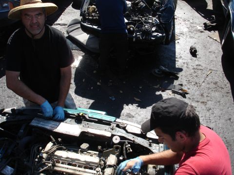 1993 Ford Taurus - Two men in a junkyard work on the engine of a car, with another vehicle's damaged front end visible in the background.
