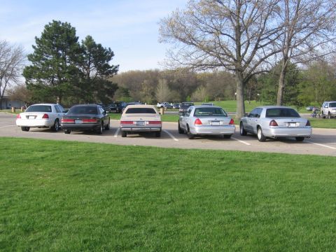 1988 Mercury Grand Marquis - Multiple sedans, likely Ford Crown Victorias, parked in a grassy lot on a clear day.