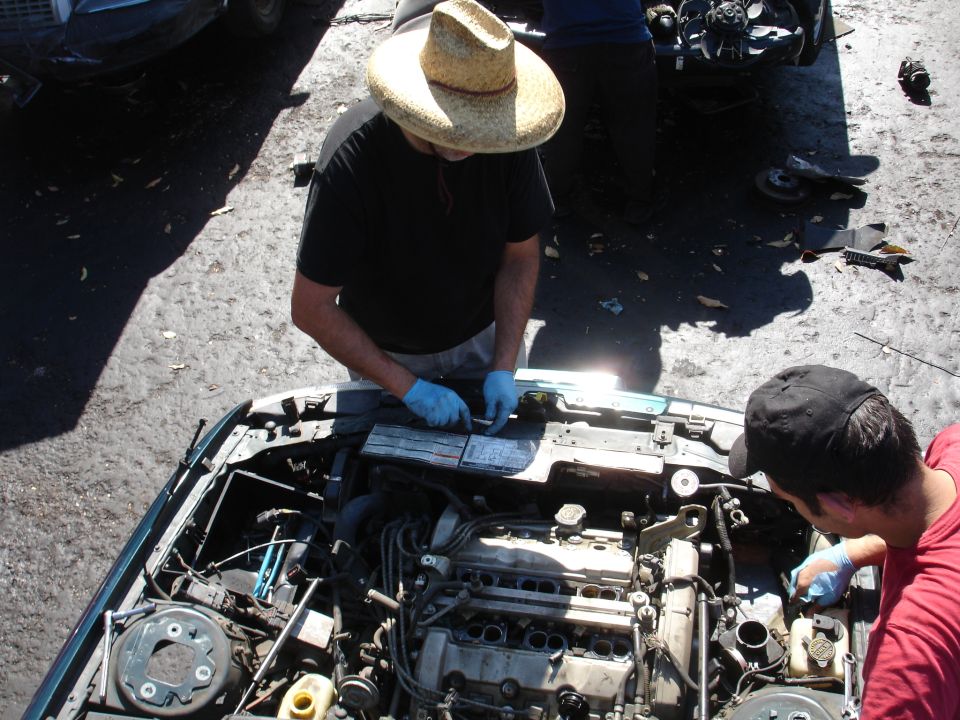 Two men in a junkyard working on a car engine, one wearing a straw hat