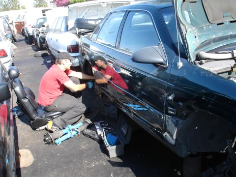 1993 Ford Taurus - A mechanic works on a dark sedan in a junkyard with other vehicles visible.