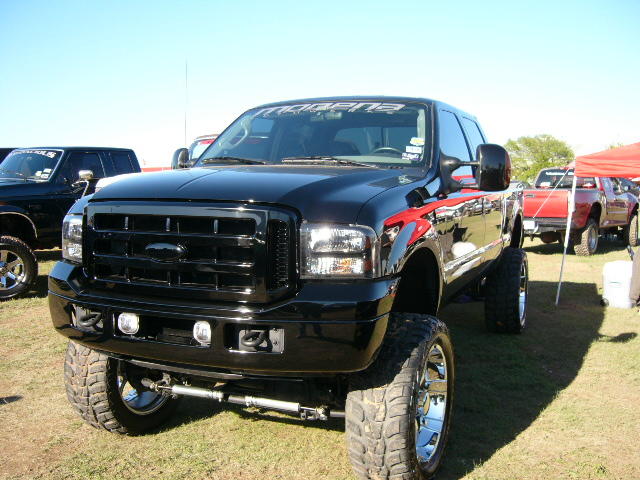 Black lifted Ford F-250 truck with chrome wheels and red graphics on the fender. Another truck and a red truck are visible in the background