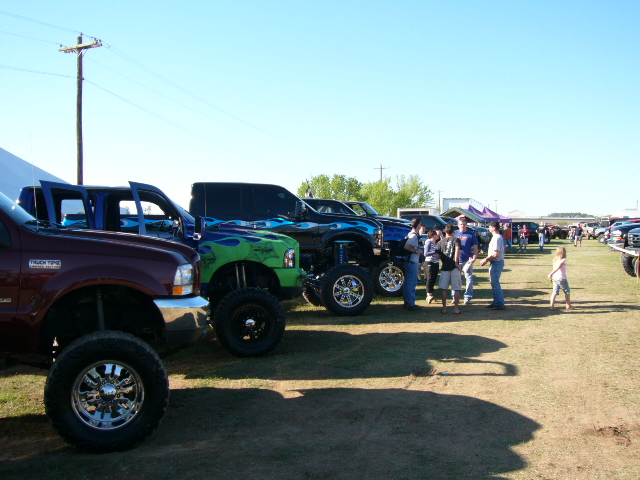 Maroon pickup truck with chrome wheels and lifted suspension, parked in a line with other modified trucks