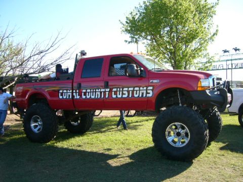 1978 Ford Bronco - Custom red Ford F-350 pickup truck with "COMAL COUNTY CUSTOMS" graphics, modified for off-road use.