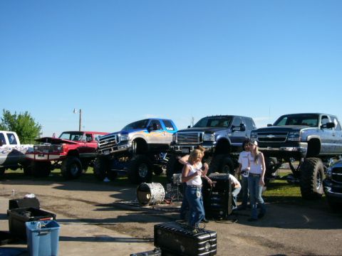 1978 Ford Bronco - A group of people stand near several large, lifted pickup trucks on a sunny day.