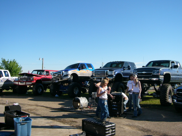Several lifted pickup trucks are parked in a lot with people standing around them. Some trucks have their hoods up