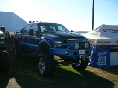 1978 Ford Bronco - A heavily modified blue Ford F-250 Super Duty truck is displayed outdoors next to vendor tents.