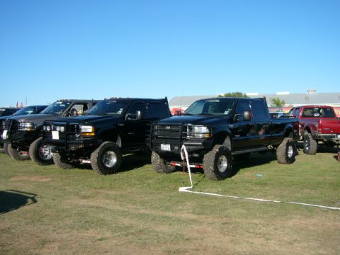 1978 Ford Bronco - Several lifted Ford trucks are parked in a line on a grassy field under a clear blue sky.