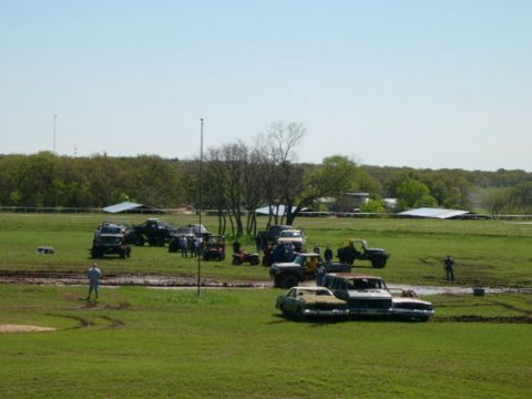1978 Ford Bronco - Pickups, ATVs and a derby car gather around a muddy pit at an open-field off-road mud bog event.