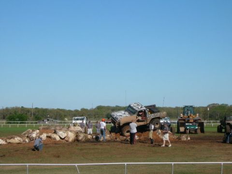 1978 Ford Bronco - Mud-covered lifted pickup climbing a pile of white boulders in a dirt obstacle course, watched by a crowd of spectators standing behind a w…